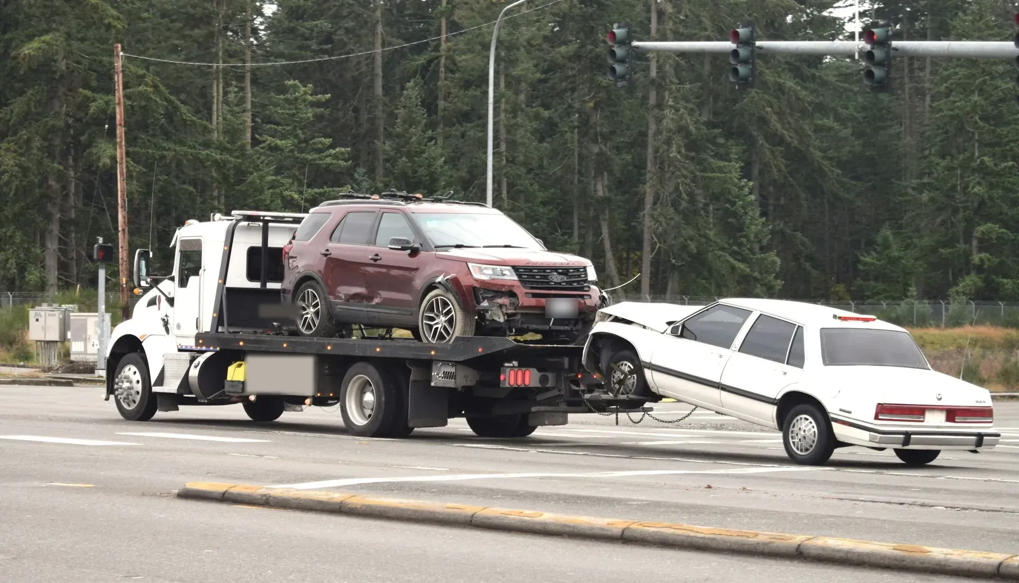 White flatbed tow truck carrying an SUV while towing a second car on a city street
